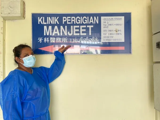 A dentist in a blue gown pointing to the Klinik Pergigian Manjeet signboard at the clinic